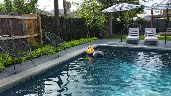 Person floating in a backyard pool beside two lounge chairs and a patio umbrella