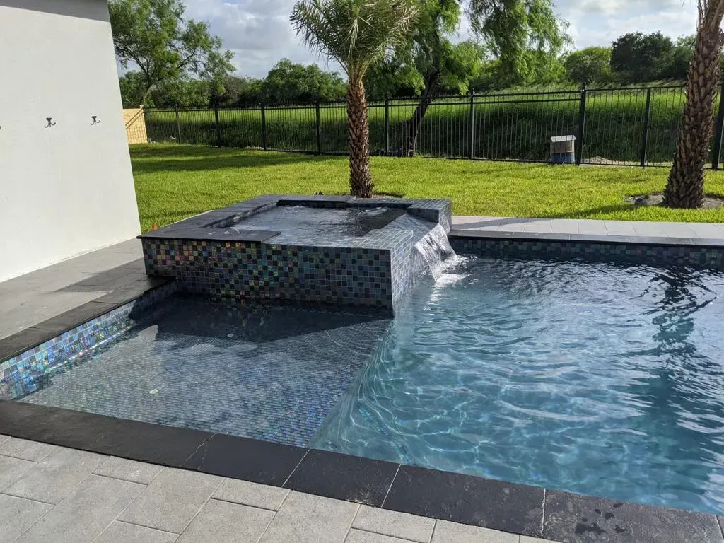Modern backyard pool with a raised waterfall feature, blue water, and green lawn beyond.
