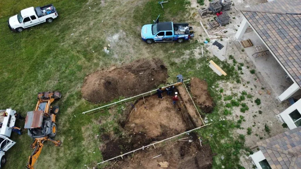 Aerial view of a yard with dirt pits, parked white and blue vehicles, and a house roof at right.