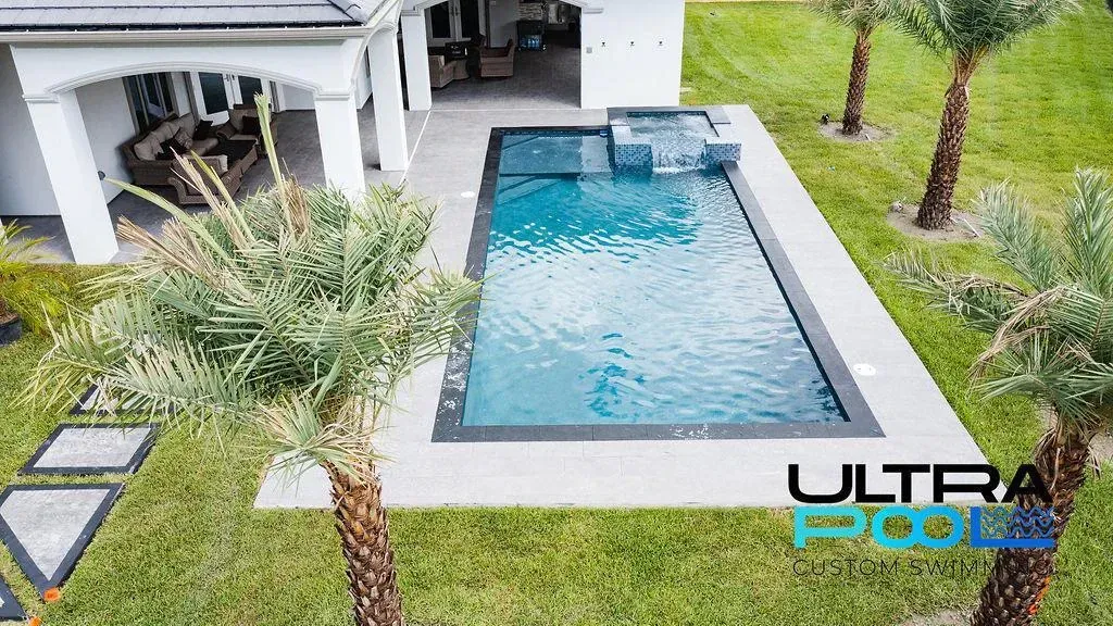 Modern backyard pool with gray deck, palm trees, and a covered patio, viewed from above