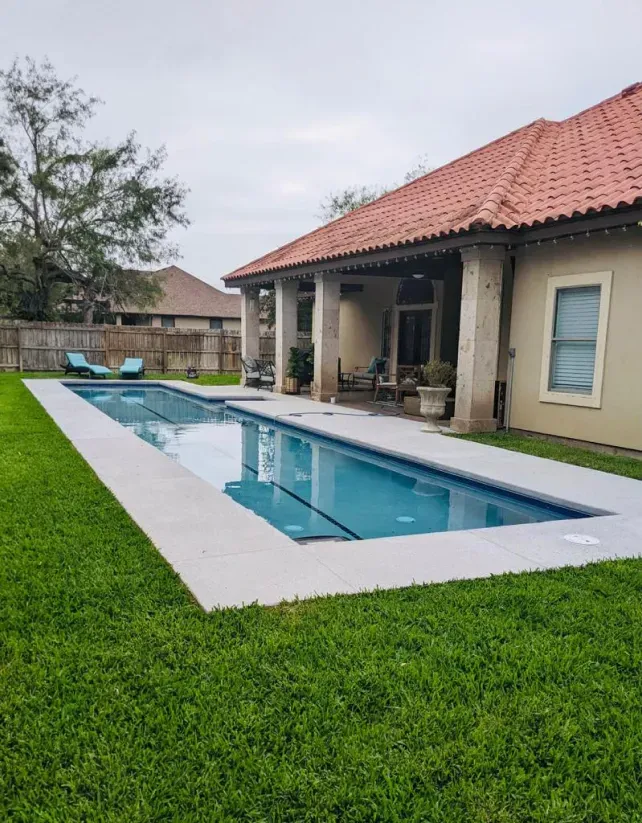 Backyard patio with a narrow pool, green lawn, and beige house with a red-tiled roof.