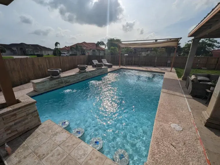 Backyard swimming pool with lounge chairs, wooden fence, and cloudy sky.
