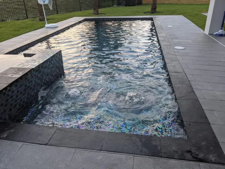 Rectangular backyard pool with rippling water, dark tile border, and gray stone patio beside a lawn