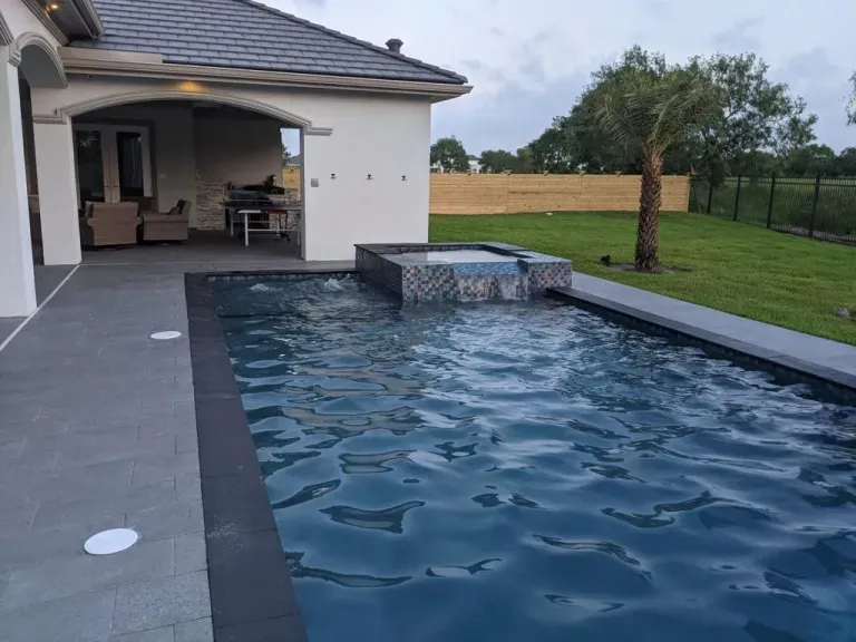 Backyard pool beside a modern house with a covered patio and green lawn at dusk