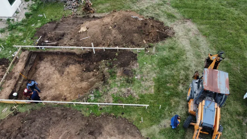 Aerial view of a rectangular excavation site with workers and a backhoe in a grassy field.