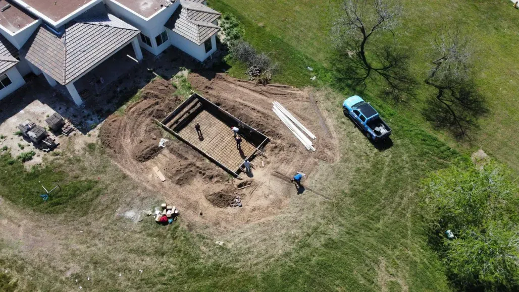Aerial view of a house under construction with a foundation, workers, and a blue truck in a grassy yard