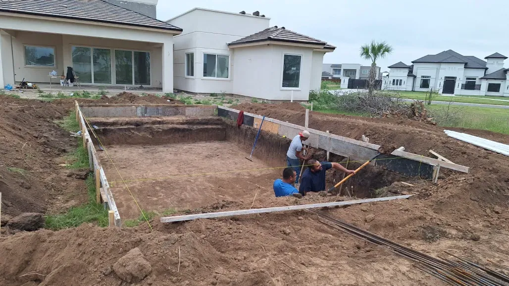 Construction crew excavating a backyard pool area beside a modern house, with dirt piles and blue equipment.