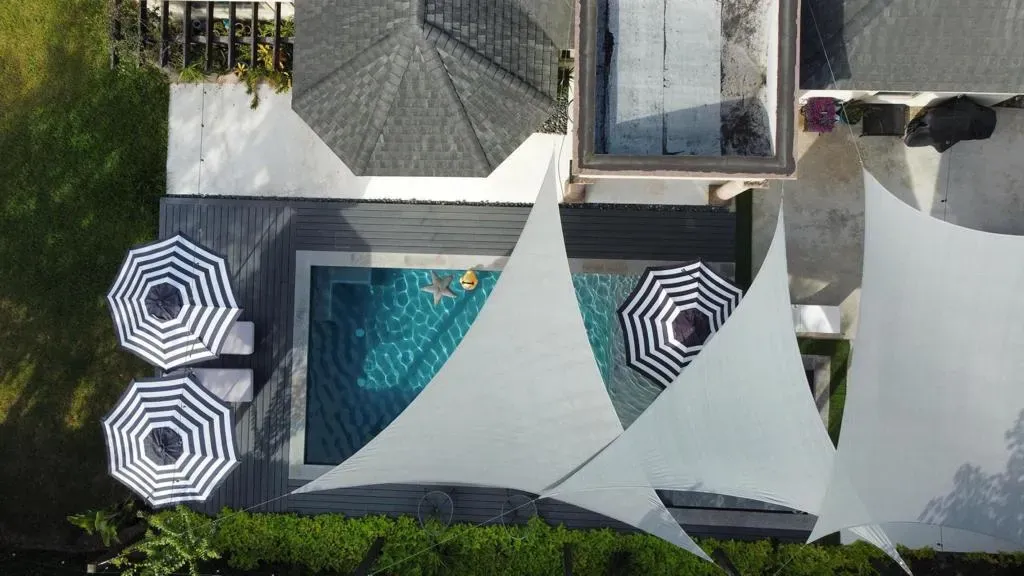 Aerial view of a backyard pool with white shade sails and striped umbrellas beside a house