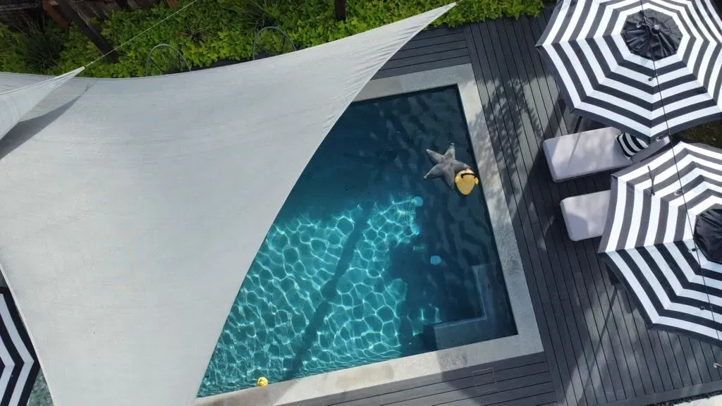 Overhead view of a pool with a person floating, striped umbrellas, and a shaded patio.