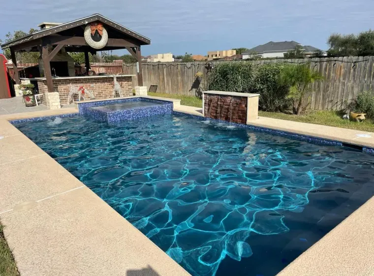 Backyard swimming pool with blue water, stone waterfall, and covered patio beside a fenced yard