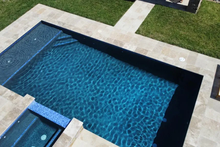 Rectangular backyard swimming pool with blue water, surrounded by light stone patio and green grass