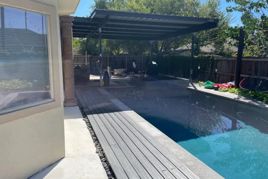 Backyard pool with covered patio and wooden deck, bright sunlight, and lounge chairs under shade