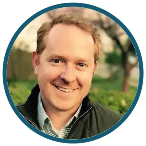 Smiling person outdoors in a circular frame, wearing a dark jacket and checkered shirt.