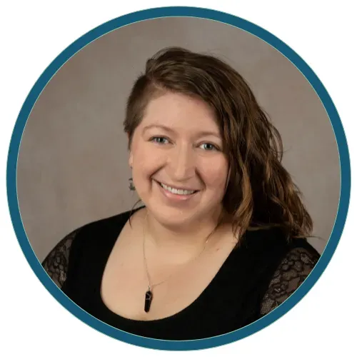 Smiling person with wavy brown hair in a black top against a gray background, framed by a teal circle