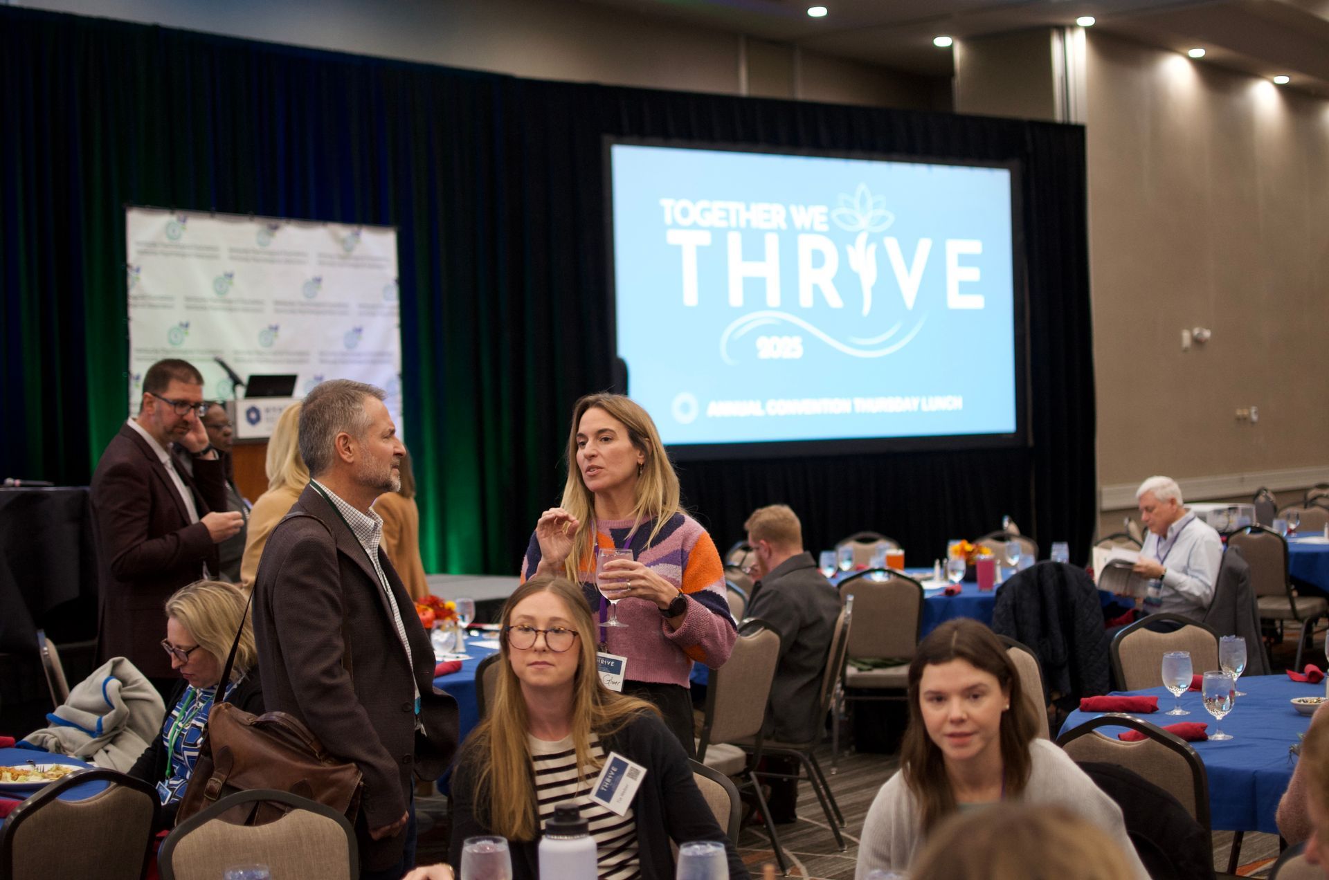 Conference attendees mingling in a hotel ballroom with a large “THRIVE” presentation screen in the background