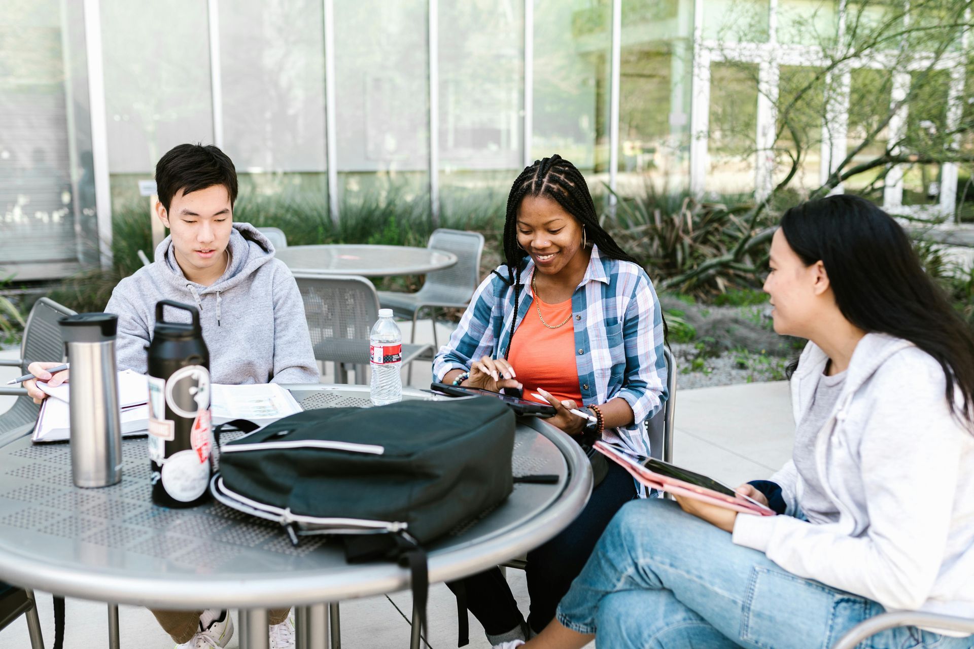 Three students sit at an outdoor café table, talking and working with notebooks and laptops.