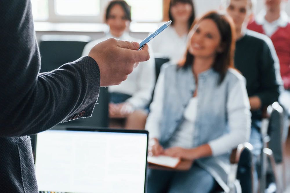 Person giving a presentation to a group in a brightly lit room. They hold a pen while looking toward the audience.