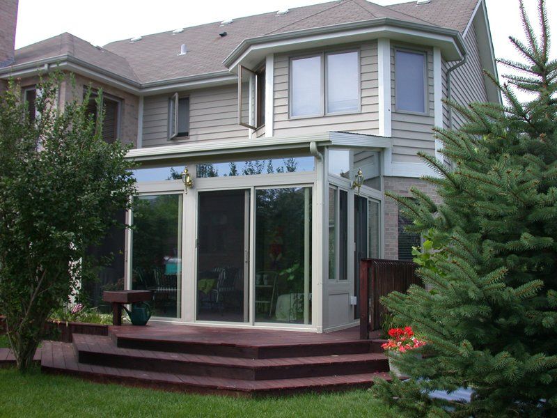 Sunroom addition on a house with a wooden deck, surrounded by trees and greenery.