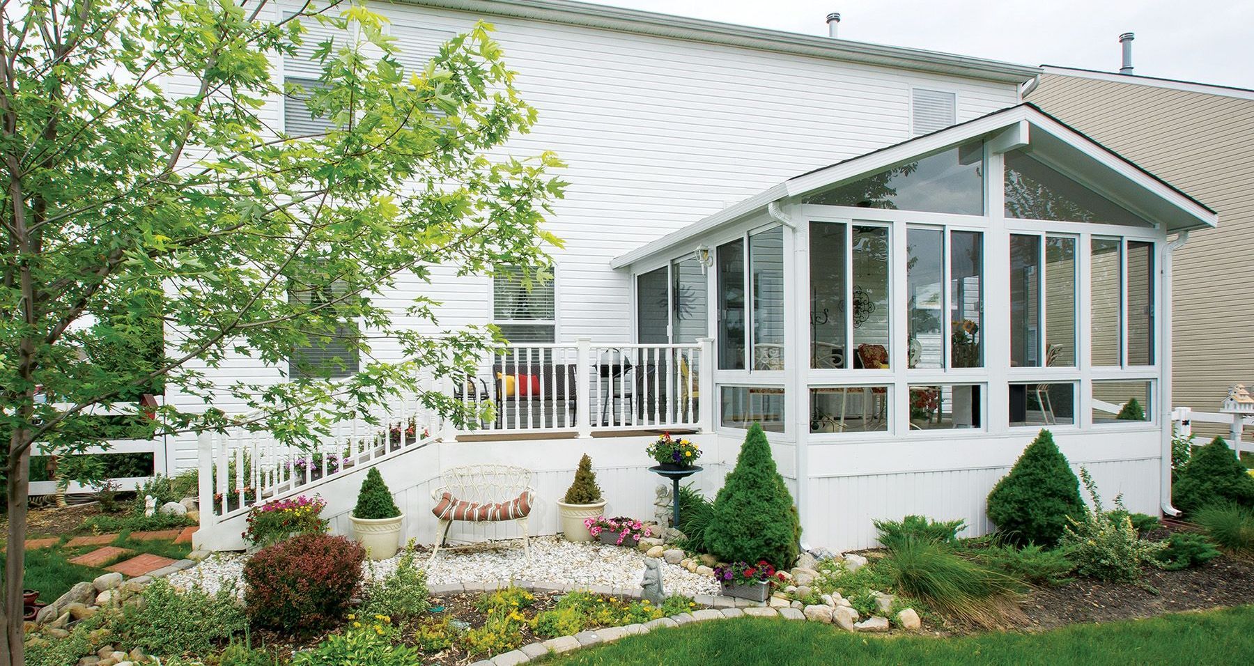 White house with a screened-in porch, deck, and garden. The porch has large windows, with a deck and white railing.