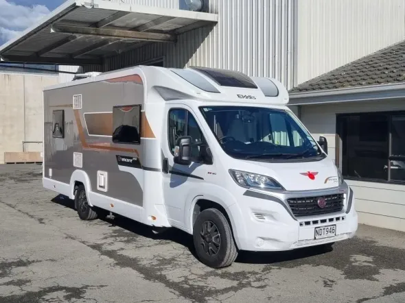 Large white-and-gray motorhome parked under a metal awning beside an industrial building