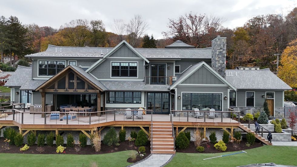 Large gray house with decks, stairs, and a screened porch overlooking a grassy yard with fall foliage in the background.