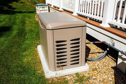 Beige outdoor generator beside a house deck, with gravel and grass around it.