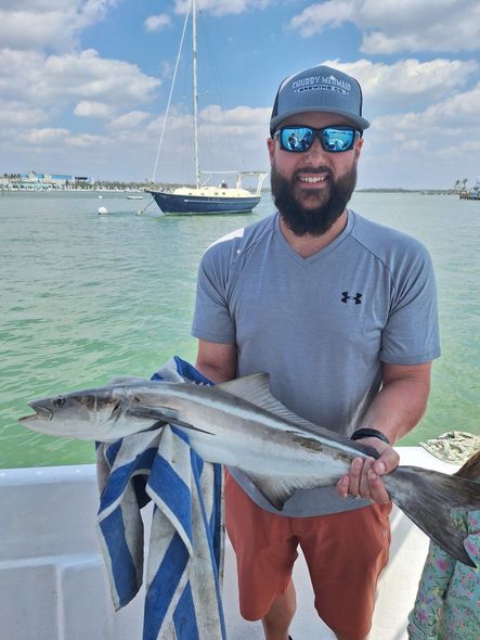 Man on boat holds up a fish he caught, smiling. Blue water, boat, harbor in background.