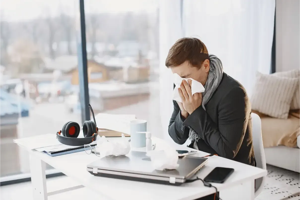 Person sneezing into a tissue at a desk with a laptop, coffee, and headphones in a bright office