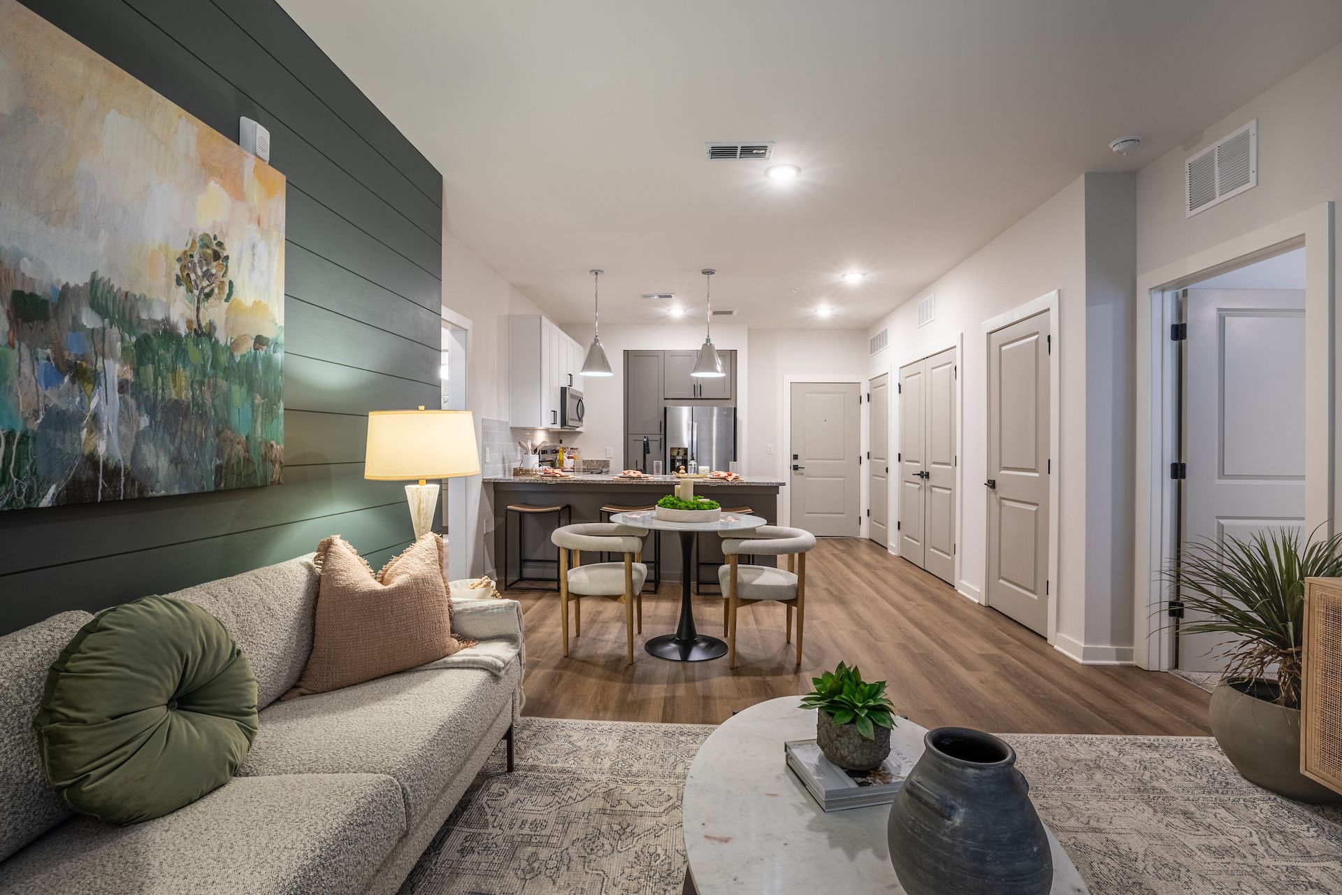 Living room with sofa, artwork, dining area, kitchen, and doorways. Light wood floors, neutral tones.