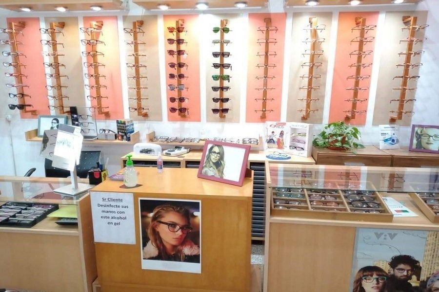Interior de una óptica con paredes de exposición de monturas, mostrador y cartel promocional enmarcado.