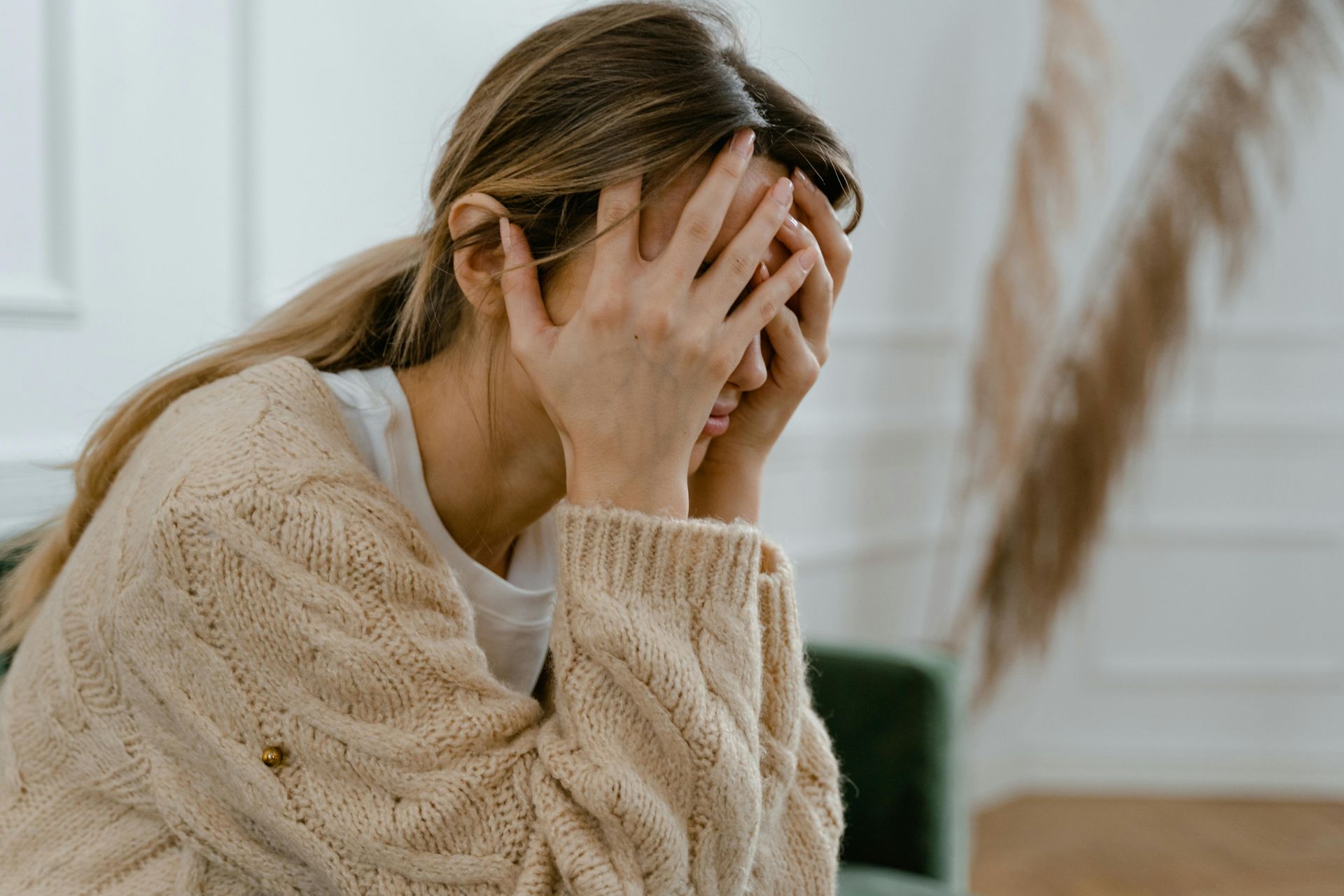 Person in beige sweater covers face with hands, sitting indoors and looking distressed