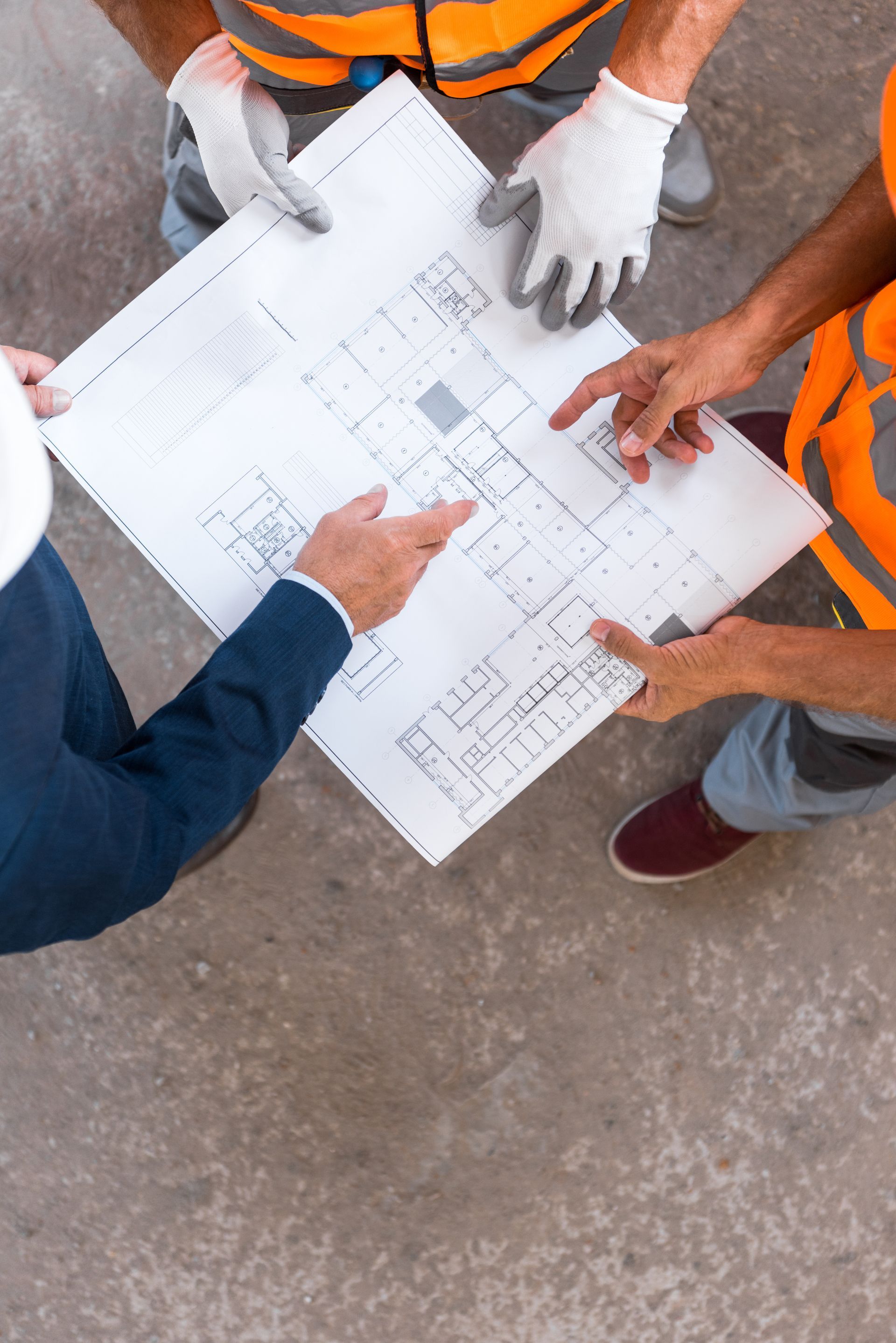 People in orange vests review blueprints on a concrete floor.