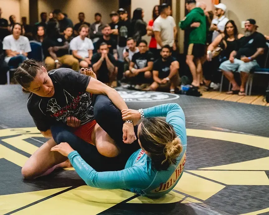 Two grapplers on a mat, one in a black shirt pinning another in a teal top, with spectators watching nearby
