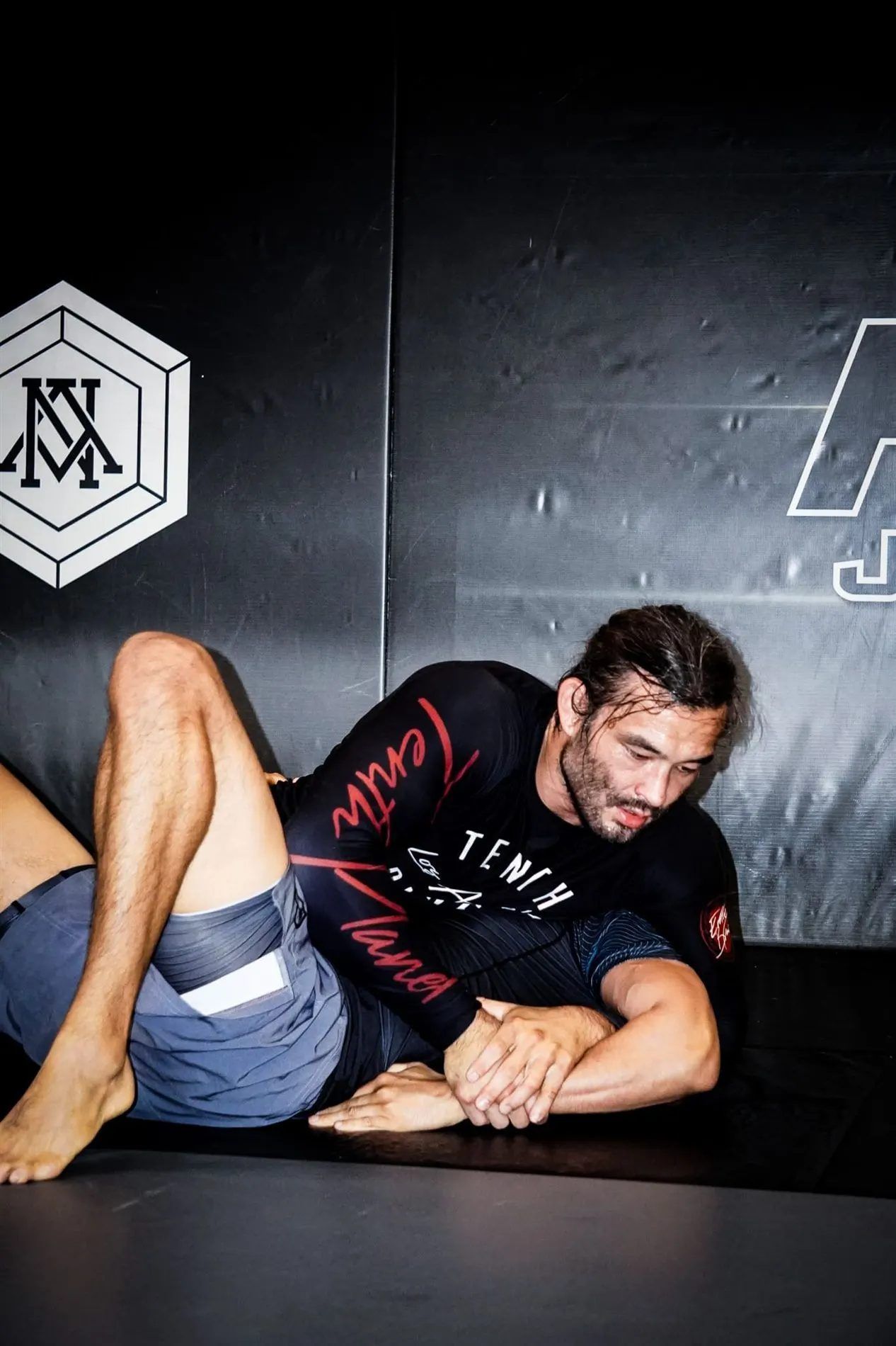 Man practicing ground grappling in a gym, wearing a black shirt and blue shorts on a mat.
