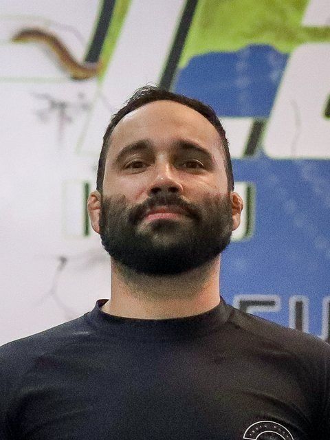 Portrait of a bearded man in a black shirt standing before a blurred blue-and-white backdrop
