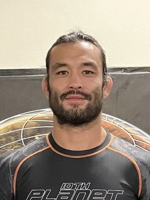 Man wearing a black athletic shirt with “10th Planet,” smiling against a gray wall.