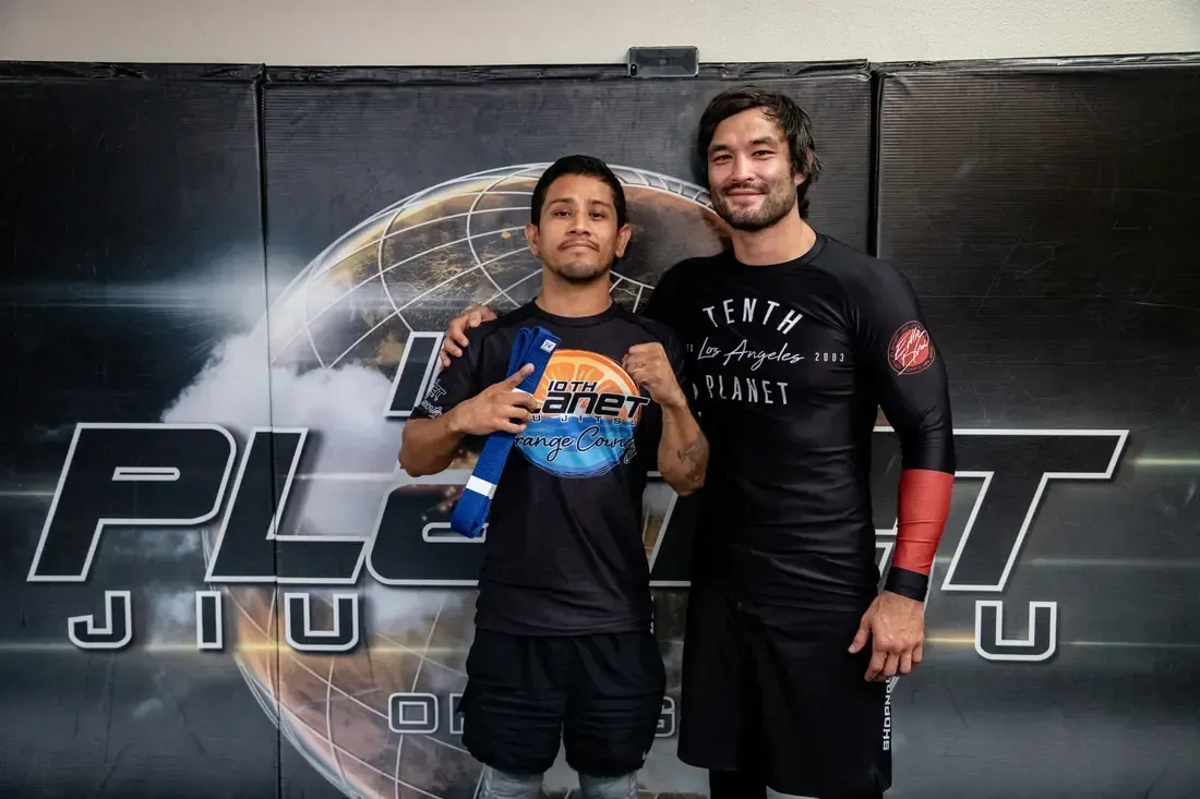 Two men in athletic gear posing in front of a large “PLO…” Jiu Jitsu banner