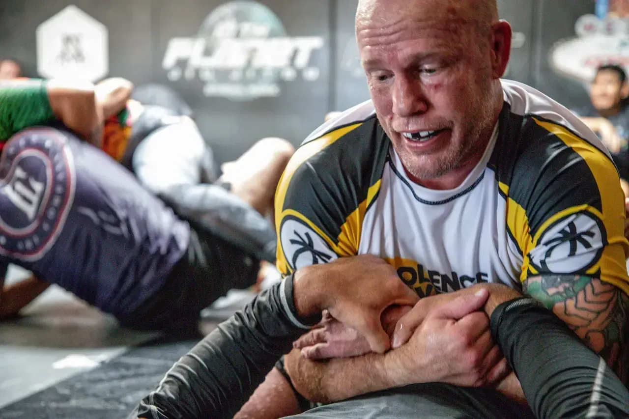 Two grapplers on mats, one in a yellow and gray rash guard applying a leg lock in a training gym