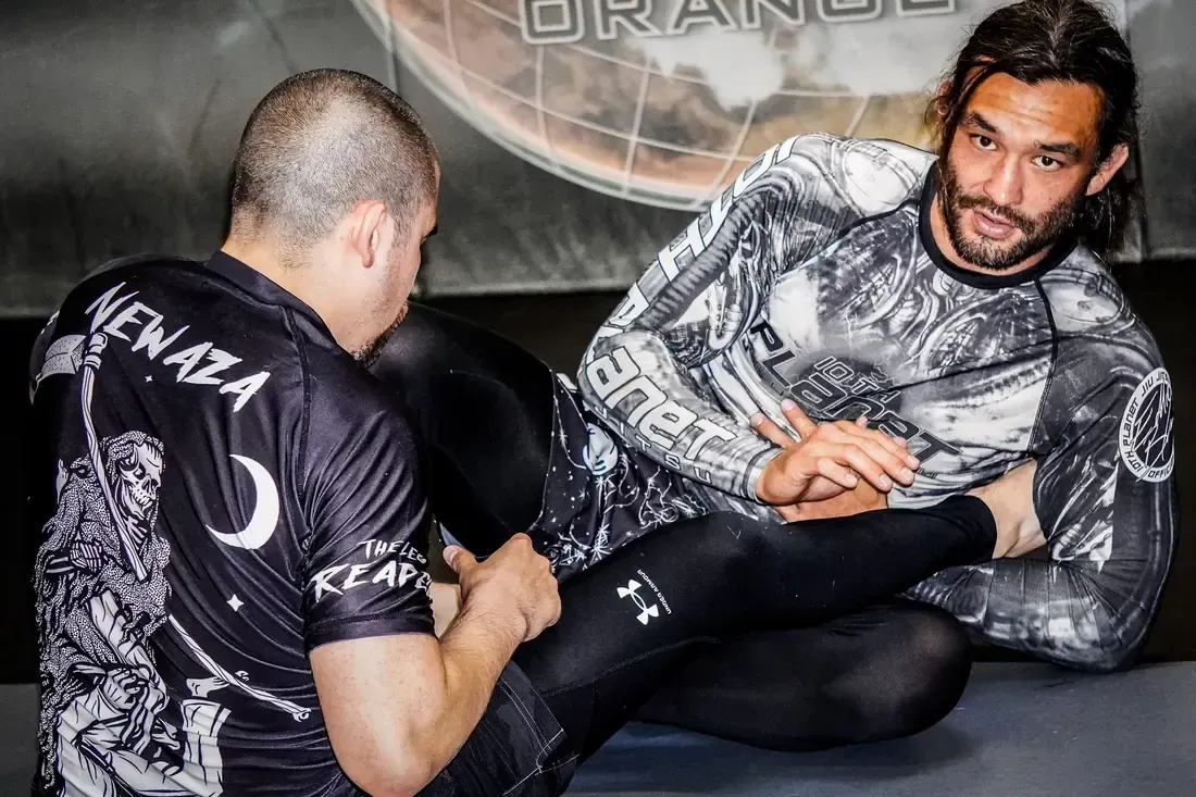 Two men grappling on a gym mat, one in black and one in gray, with a training logo in the background