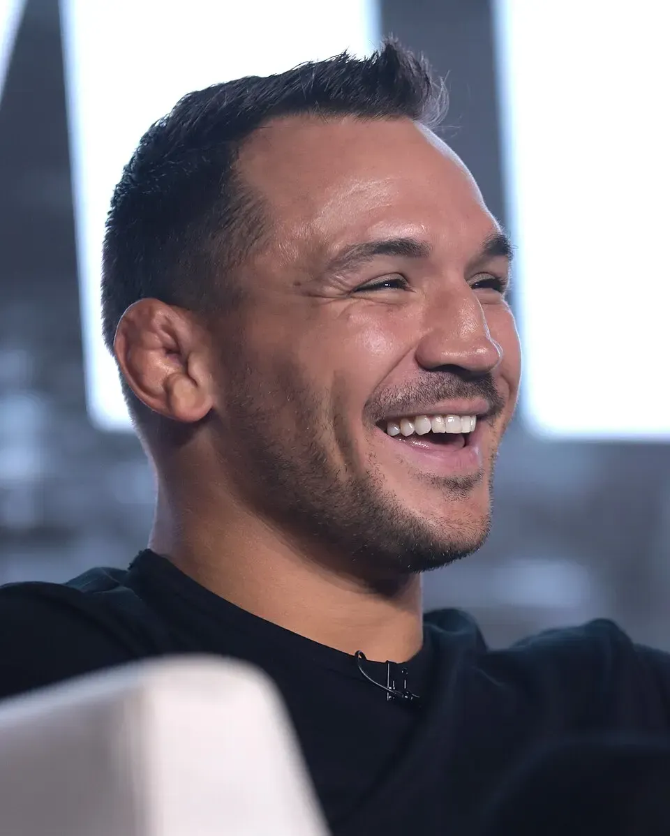 Smiling man in a black shirt, seated indoors with a blurred bright background.