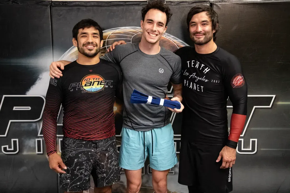 Three men in athletic gear pose smiling in a gym, standing in front of a black wall with large lettering.