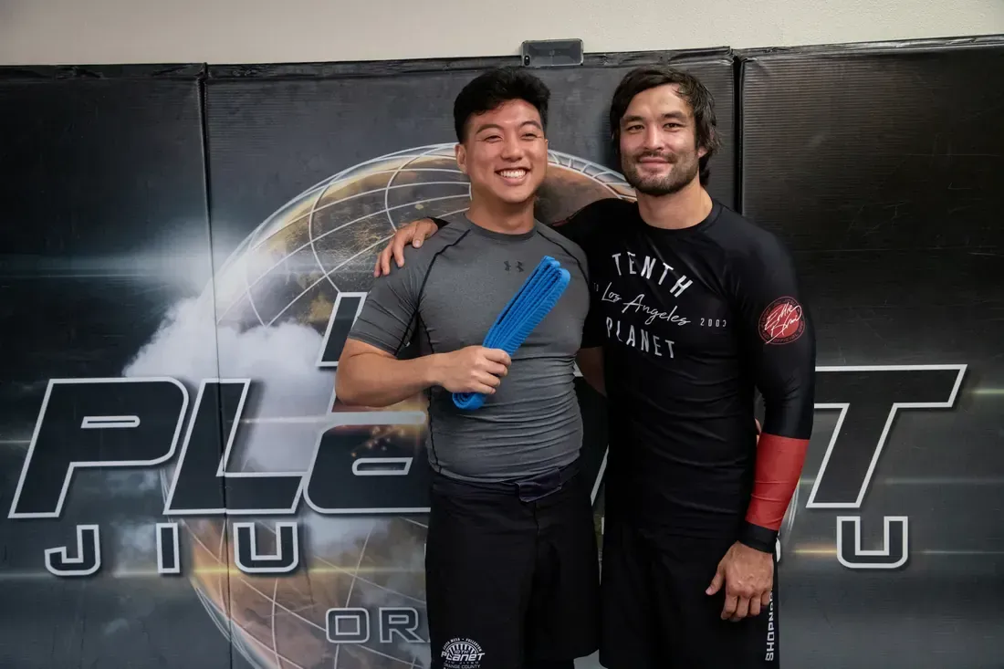 Two smiling grapplers pose in front of a Planet Jiu-Jitsu banner after training.