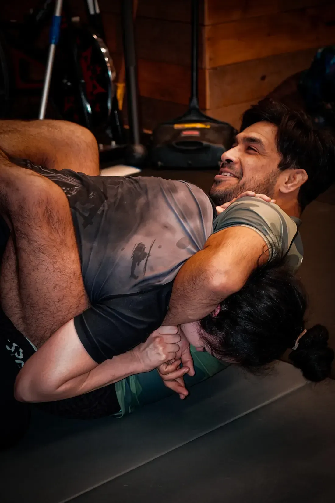 Two wrestlers grappling on a mat in a gym, one in a gray shirt and black shorts.
