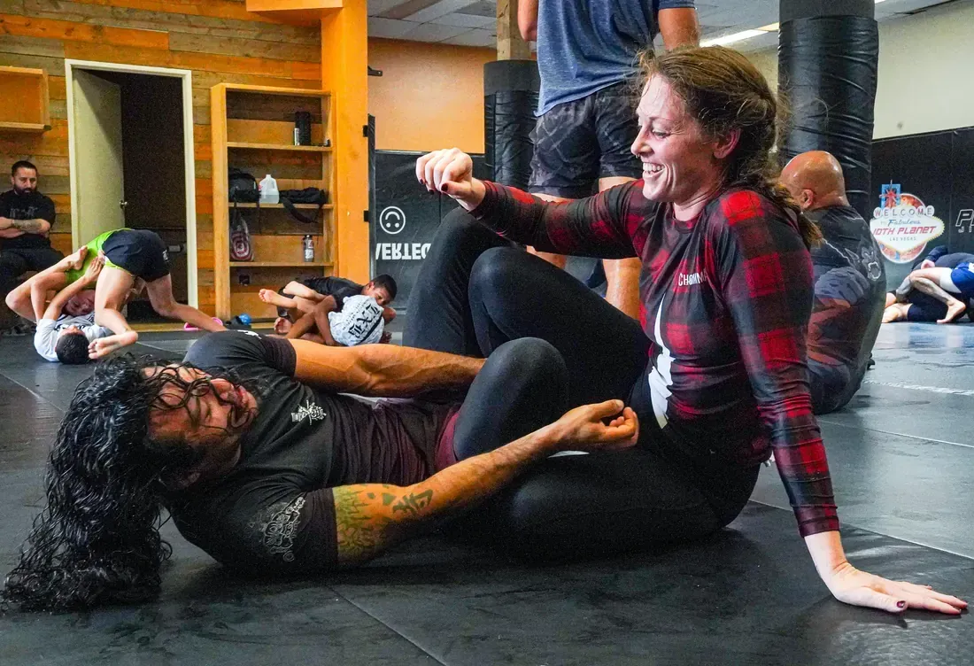 Two people grappling on gym mats while others train in the background