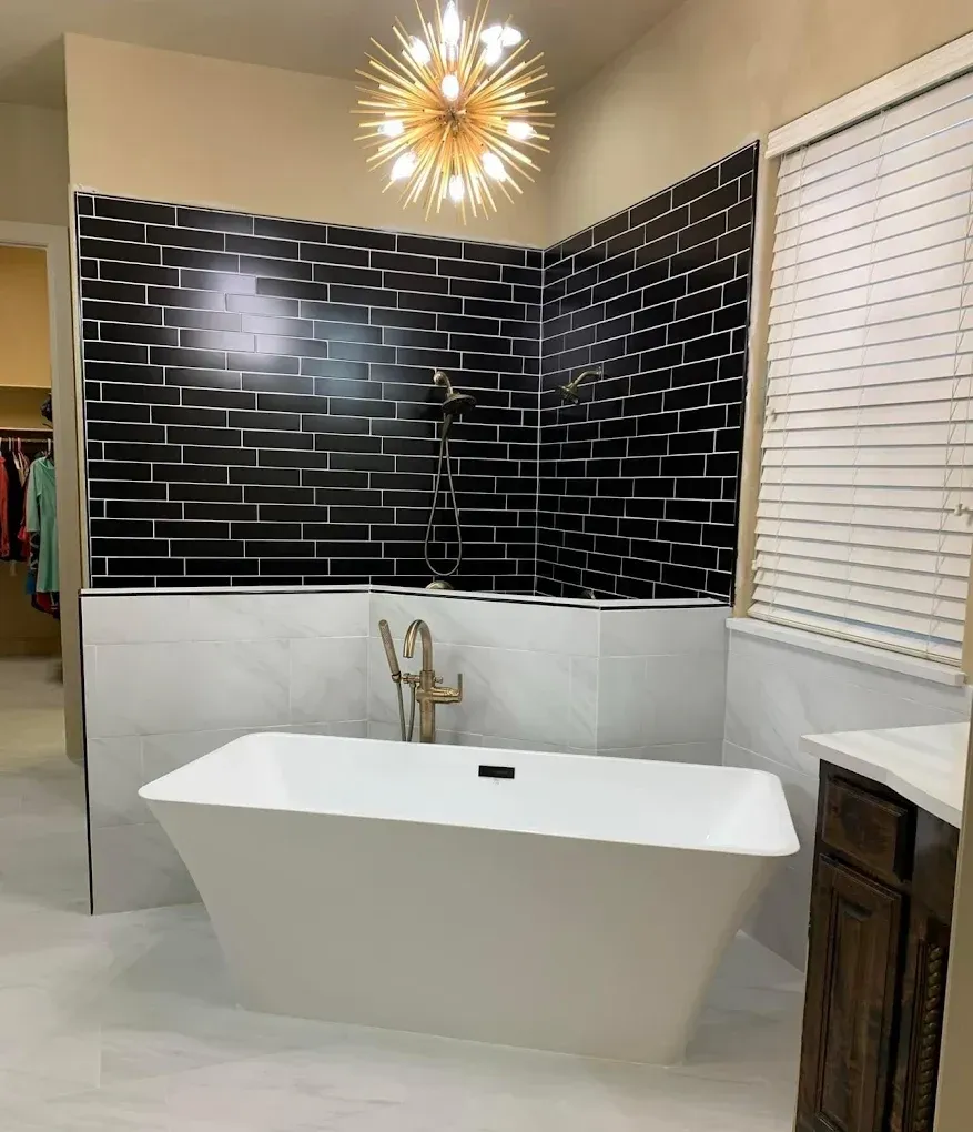 Modern bathroom with black brick-like tile, white tub, gold fixtures, and a chandelier.