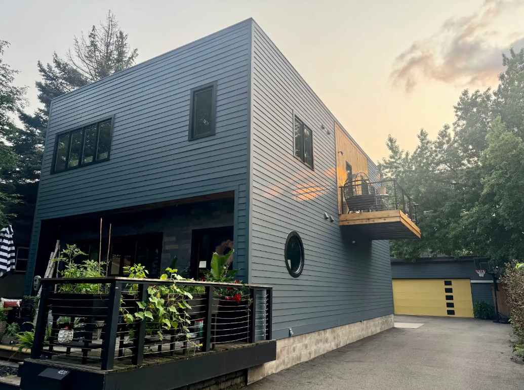 Modern two-story house with dark gray exterior, small balcony, and yellow garage. Surrounded by trees, overcast sky.