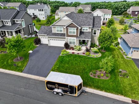 Two-story house with a trailer parked in the street. Green lawn, trees, and other houses in the background.