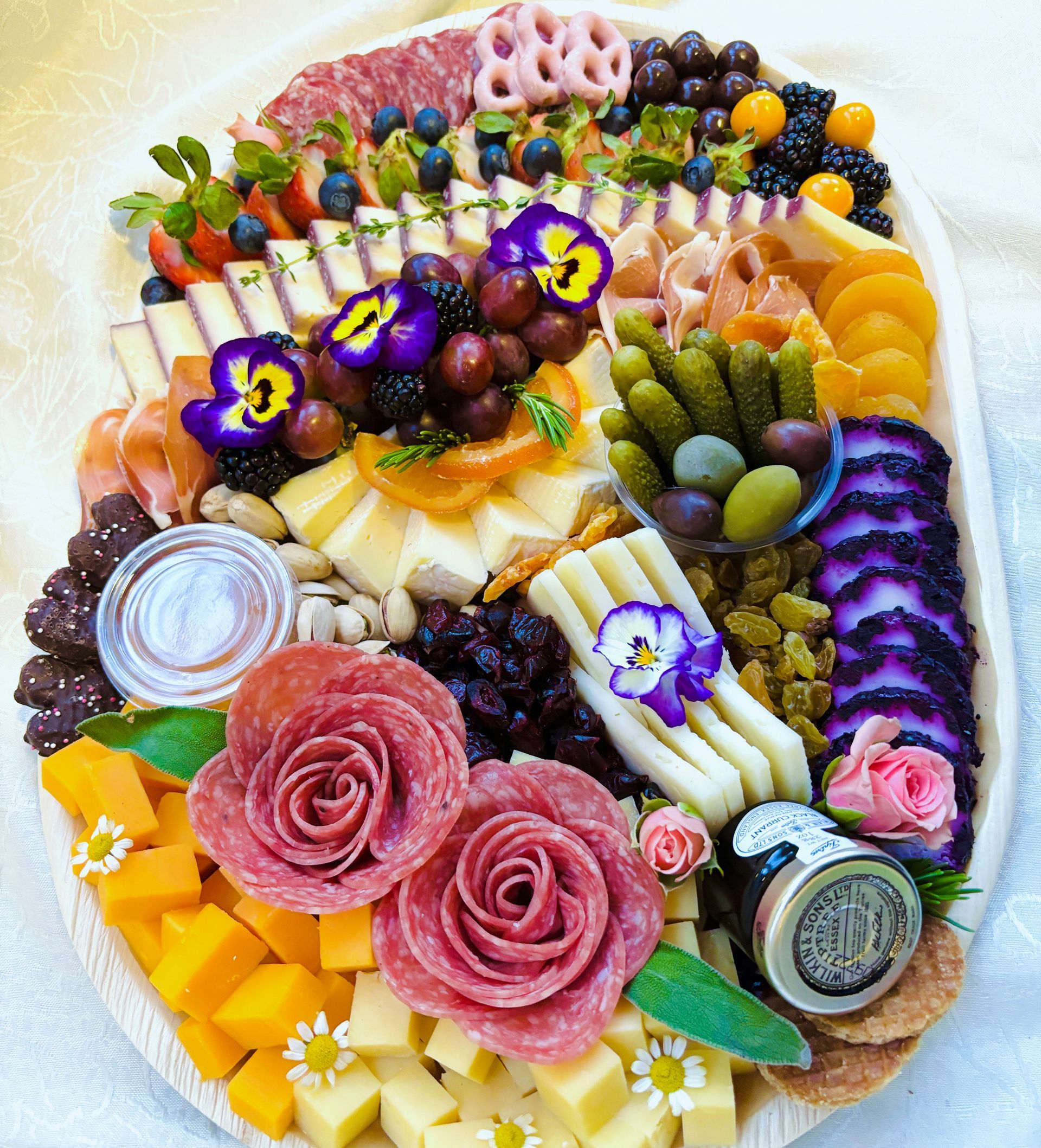 Oval charcuterie board with cheese, meats, fruits, nuts, and flowers arranged decoratively.