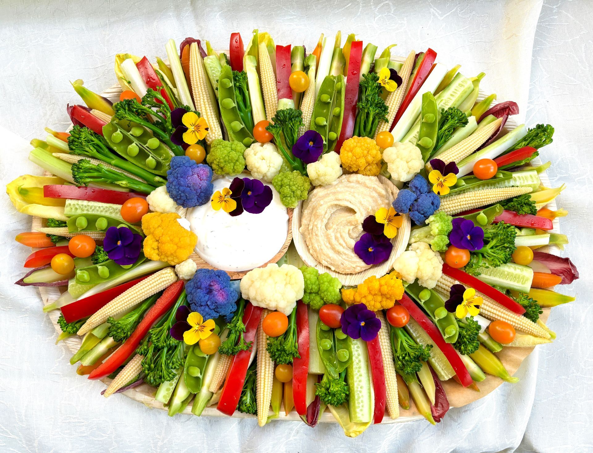 Vegetable platter with two dips, colorful arrangement of raw vegetables and edible flowers on a white surface.