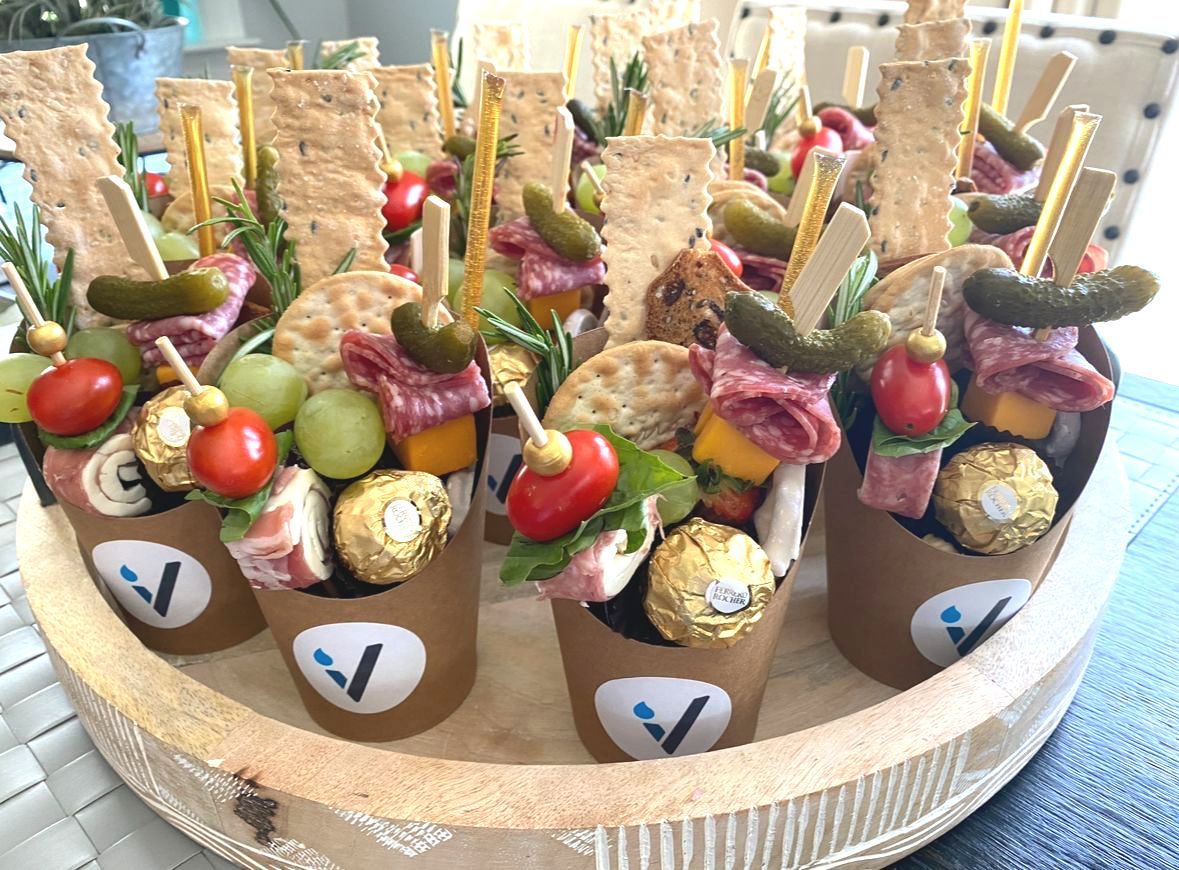 Appetizer cups with crackers, fruit, pickles, meat, cheese, and chocolate on a wooden tray.